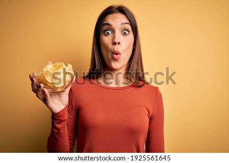 Young beautiful brunette woman holding bowl with chips potatoes over yellow background scared in shock with a surprise face, afraid and excited with fear expression