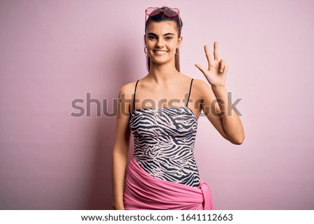 Young beautiful brunette woman on vacation wearing swimsuit over pink background showing and pointing up with fingers number three while smiling confident and happy.