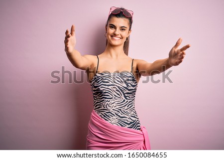 Young beautiful brunette woman on vacation wearing swimsuit over pink background looking at the camera smiling with open arms for hug. Cheerful expression embracing happiness.