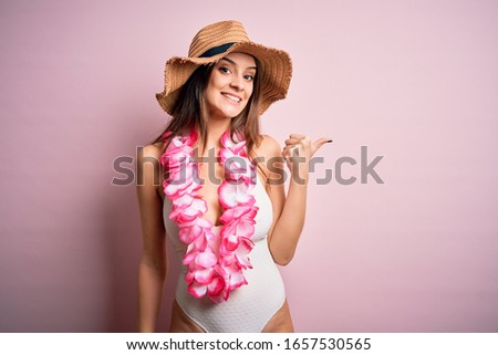 Young beautiful brunette woman on vacation wearing swimsuit and Hawaiian flowers lei smiling with happy face looking and pointing to the side with thumb up.