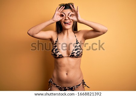 Young beautiful brunette woman on vacation wearing swimwear bikini over yellow background doing ok gesture like binoculars sticking tongue out, eyes looking through fingers. Crazy expression.