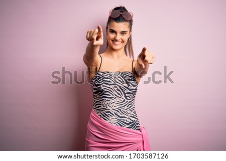 Young beautiful brunette woman on vacation wearing swimsuit over pink background pointing to you and the camera with fingers, smiling positive and cheerful