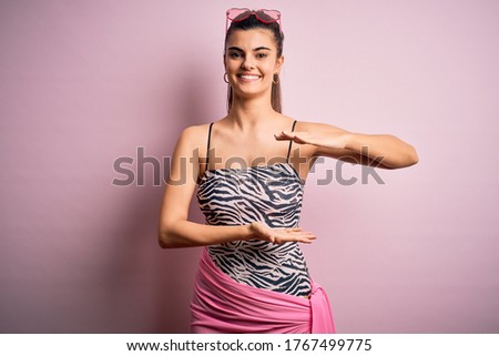 Young beautiful brunette woman on vacation wearing swimsuit over pink background gesturing with hands showing big and large size sign, measure symbol. Smiling looking at the camera. Measuring concept.