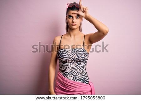 Young beautiful brunette woman on vacation wearing swimsuit over pink background making fun of people with fingers on forehead doing loser gesture mocking and insulting.