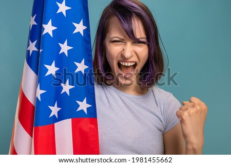 Young Beautiful Brunette Woman With Purple Dyed Hair And Piercings With American Stars Stripes USA Flag In Her Hands On A Blue Background