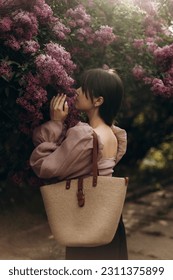 young beautiful brunette woman reaches for the branches of blooming lilacs. a girl in a beige blouse with a straw bag near a blooming lilac. - Shutterstock ID 2311375899