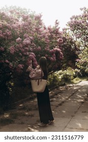 young beautiful brunette woman reaches for the branches of blooming lilacs. a girl in a beige blouse with a straw bag near a blooming lilac. - Shutterstock ID 2311516779