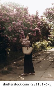 young beautiful brunette woman reaches for the branches of blooming lilacs. a girl in a beige blouse with a straw bag near a blooming lilac. - Shutterstock ID 2311517675