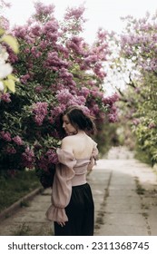 young beautiful brunette woman stands with her back near a blooming lilac. a girl in a beige blouse with an open back stands in a lilac garden - Shutterstock ID 2311368745
