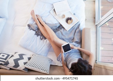 Young Beautiful Brunette Woman With Tablet Computer  Sitting Down On The Bed By The Window. Top View From Above. Lazy Day Off Concept