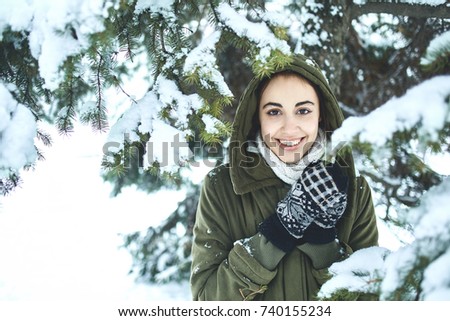 young beautiful brunette woman in a warm jacket with hoodie and warm mittens is walking outdoors in winter. a woman stands under the fir branches with snow and play with them
