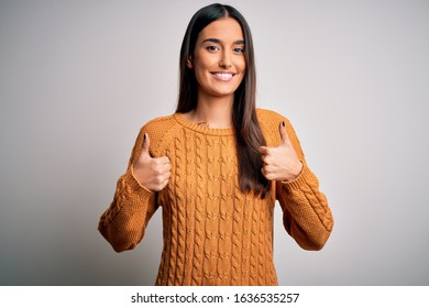 Young beautiful brunette woman wearing casual sweater over isolated white background success sign doing positive gesture with hand, thumbs up smiling and happy. Cheerful expression and winner gesture.