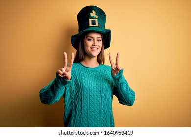 Young beautiful brunette woman wearing green hat with clover celebrating saint patricks day smiling looking to the camera showing fingers doing victory sign. Number two.