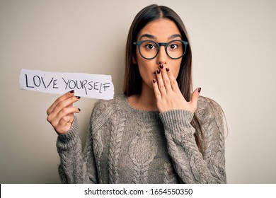 Young beautiful brunette woman wearing glasses holding paper with love yourself message cover mouth with hand shocked with shame for mistake, expression of fear, scared in silence, secret concept