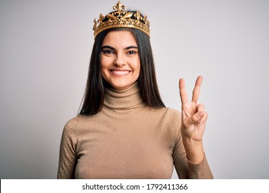 Young beautiful brunette woman wearing golden queen crown over isolated white background smiling with happy face winking at the camera doing victory sign. Number two.