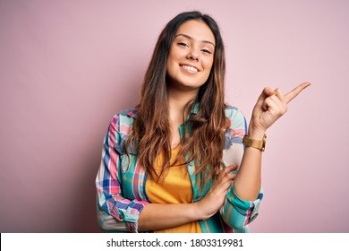 Young beautiful brunette woman wearing casual colorful shirt standing over pink background with a big smile on face, pointing with hand and finger to the side looking at the camera.