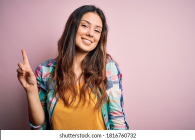 Young beautiful brunette woman wearing casual colorful shirt standing over pink background showing and pointing up with finger number one while smiling confident and happy.