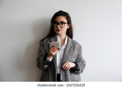 Young Beautiful Brunette Woman Wearing Smart Casual Attire, Posing With A Phone Over White Wall Background. Portrait Of Female Model Typing On Her Cellphone. Close Up, Copy Space.