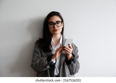 Young Beautiful Brunette Woman Wearing Smart Casual Attire, Posing With A Phone Over White Wall Background. Portrait Of Female Model Typing On Her Cellphone. Close Up, Copy Space.