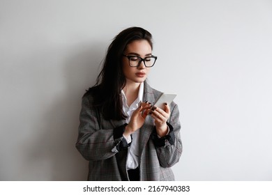 Young Beautiful Brunette Woman Wearing Smart Casual Attire, Posing With A Phone Over White Wall Background. Portrait Of Female Model Typing On Her Cellphone. Close Up, Copy Space.