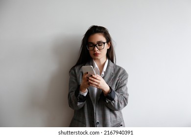 Young Beautiful Brunette Woman Wearing Smart Casual Attire, Posing With A Phone Over White Wall Background. Portrait Of Female Model Typing On Her Cellphone. Close Up, Copy Space.