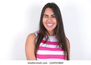 Young Beautiful Brunette Woman Wearing Striped Crop Top Over White Wall Blinking Eyes With Pleasure Having Happy Expression. Facial Expressions And People Emotions Concept.