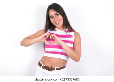 Young beautiful brunette woman wearing striped crop top over white wall smiling in love showing heart symbol and shape with hands. Romantic concept.