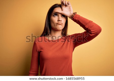 Young beautiful brunette woman wearing casual t-shirt standing over yellow background making fun of people with fingers on forehead doing loser gesture mocking and insulting.