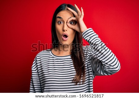 Young beautiful brunette woman wearing casual striped t-shirt over red background doing ok gesture shocked with surprised face, eye looking through fingers. Unbelieving expression.