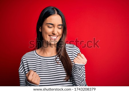 Young beautiful brunette woman wearing casual striped t-shirt over red background excited for success with arms raised and eyes closed celebrating victory smiling. Winner concept.