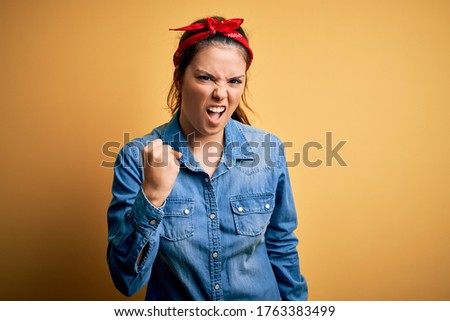 Young beautiful brunette woman wearing casual denim shirt and hair handkerchief angry and mad raising fist frustrated and furious while shouting with anger. Rage and aggressive concept.
