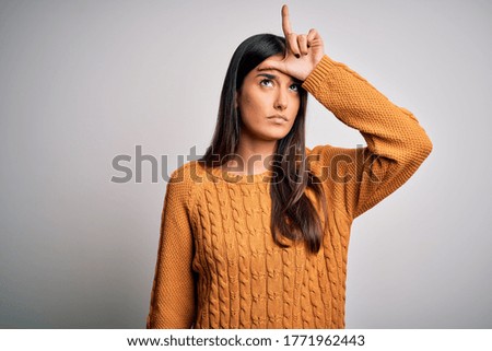 Young beautiful brunette woman wearing casual sweater over isolated white background making fun of people with fingers on forehead doing loser gesture mocking and insulting.