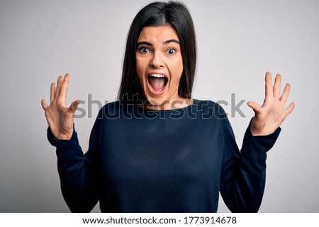 Young beautiful brunette woman wearing casual sweater standing over white background celebrating crazy and amazed for success with arms raised and open eyes screaming excited. Winner concept
