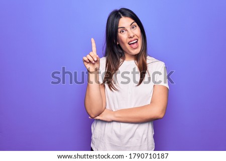 Young beautiful brunette woman wearing casual t-shirt standing over purple background smiling with an idea or question pointing finger up with happy face, number one