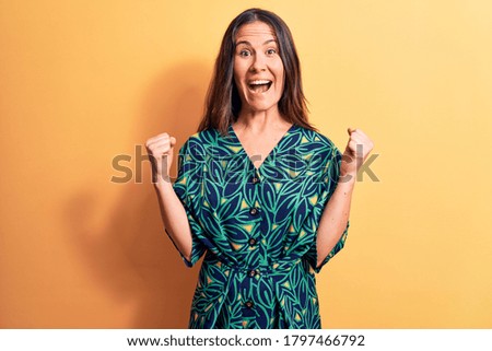 Young beautiful brunette woman wearing casual floral dress standing over yellow background celebrating surprised and amazed for success with arms raised and open eyes. Winner concept.