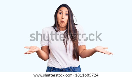 Young beautiful brunette woman wearing casual t-shirt clueless and confused expression with arms and hands raised. doubt concept. 