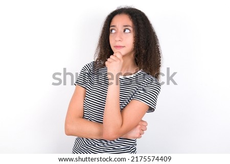 young beautiful brunette woman wearing striped t-shirt over white wall with hand under chin and looking sideways with doubtful and skeptical expression, suspect and doubt.
