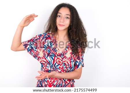 young beautiful brunette woman wearing colourful dress over white wall gesturing with hands showing big and large size sign, measure symbol.