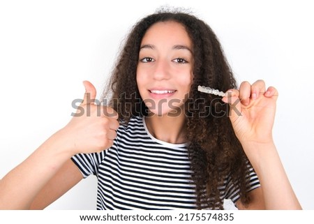young beautiful brunette woman wearing striped t-shirt over white wall holding an invisible braces aligner and rising thumb up, recommending this new treatment. Dental healthcare concept.