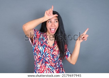 young beautiful brunette woman wearing colourful dress over white wall showing loser sign and pointing at empty space
