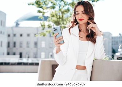 Young beautiful brunette woman in  white suit jacket.  Smiling model posing in the street . Fashionable female outdoors. Holds smartphone, uses phone apps, looks at screen, red lips - Powered by Shutterstock