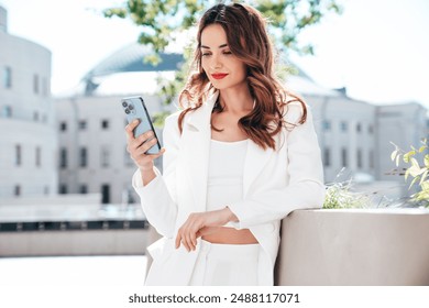 Young beautiful brunette woman in  white suit jacket.  Smiling model posing in the street . Fashionable female outdoors. Holds smartphone, uses phone apps, looks at cellphone  screen, red lips - Powered by Shutterstock