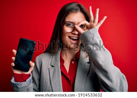Young beautiful business woman with blue eyes holding smartphone showing screen with happy face smiling doing ok sign with hand on eye looking through fingers