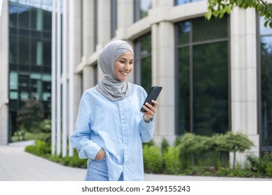 A young beautiful business woman in a hijab walks through the city, a Muslim woman holds a phone in her hands, uses an application on a smartphone, smiles, browses the Internet. - Powered by Shutterstock