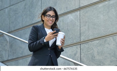 Young beautiful business woman (student) in suit, smiling, happy, walking down stairs, steps, on building background, coffee in plastic cup. Concept: new business, communication, Arab, banker, glasses