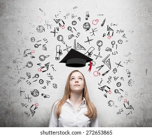 Young beautiful business woman is thinking about education at business school. Drawn business icons over the concrete wall. Graduation hat. - Shutterstock ID 272653865
