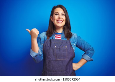 Young beautiful business woman wearing store uniform apron over blue isolated background smiling with happy face looking and pointing to the side with thumb up.