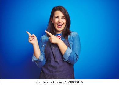 Young beautiful business woman wearing store uniform apron over blue isolated background smiling and looking at the camera pointing with two hands and fingers to the side.