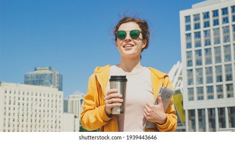 Young beautiful business woman wearing sunglasses and yellow hoodie is walking down city street, holding coffee in cup thermo mug and tablet computer. Building background.