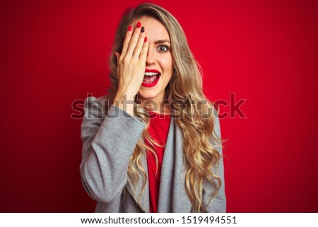 Young beautiful business woman wearing elegant jacket standing over red isolated background covering one eye with hand, confident smile on face and surprise emotion.